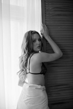 Uzun kıvırcık saçlı, iç çamaşırlı, yatak odasının penceresinin yanındaki battaniyeye sarılmış genç ve çekici bir kız.