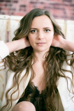 Uzun kıvırcık saçlı, dantelli siyah ceketli ve beyaz, şık ceketli çekici bir kadın stüdyoda yerde oturuyor.