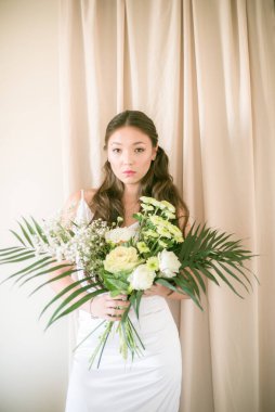 Stüdyoda bir buket çiçekli, beyaz elbiseli, uzun saçlı, Asyalı tatlı genç gelin.