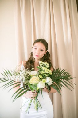 Stüdyoda bir buket çiçekli, beyaz elbiseli, uzun saçlı, Asyalı tatlı genç gelin.