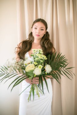 Stüdyoda bir buket çiçekli, beyaz elbiseli, uzun saçlı, Asyalı tatlı genç gelin.