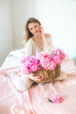 Sarışın, beyaz elbiseli, dantelli iç çamaşırlı ve stüdyodaki yatağın üzerinde pembe şakayık sepeti olan sevimli genç bir kız. Bahar ve çiçekler. Güzellik ve Moda