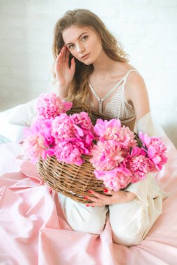Sarışın, beyaz elbiseli, dantelli iç çamaşırlı ve stüdyodaki yatağın üzerinde pembe şakayık sepeti olan sevimli genç bir kız. Bahar ve çiçekler. Güzellik ve Moda
