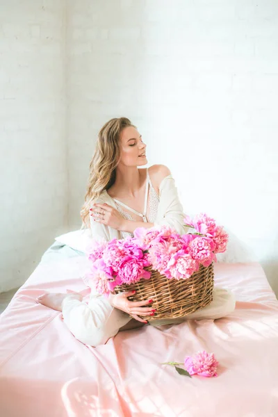 在演播室里 漂亮的年轻姑娘 金发碧眼 穿着白衣 穿着花边内裤 床上挂着一篮粉红色的牡丹 春天和花朵 美与时尚 — 图库照片