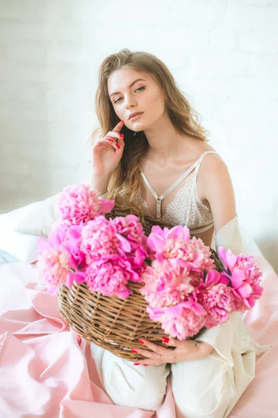 在演播室里 漂亮的年轻姑娘 金发碧眼 穿着白衣 穿着花边内裤 床上挂着一篮粉红色的牡丹 春天和花朵 美与时尚 — 图库照片
