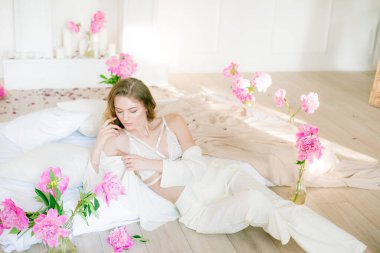Beyaz elbiseli, dantelli iç çamaşırlı, sarı saçlı sevimli genç kız. Stüdyodaki yatağın üzerinde pembe şakayıklarla çevrili. Bahar ve çiçekler. Güzellik ve Moda
