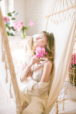 Etrafı pembe şakayıklarla çevrili, sarı saçlı, sevimli genç kız stüdyoda bir hamakta dinleniyor. Bahar ve çiçekler. Güzellik ve Moda