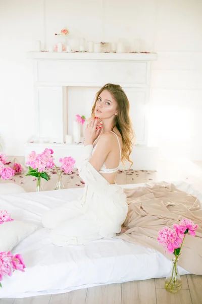 スタジオのベッドの上のベッドの上にピンクの牡丹に囲まれた白い服とレースの下着で金髪の髪のかわいい柔らかい若い女の子 春と花 美とファッション — ストック写真