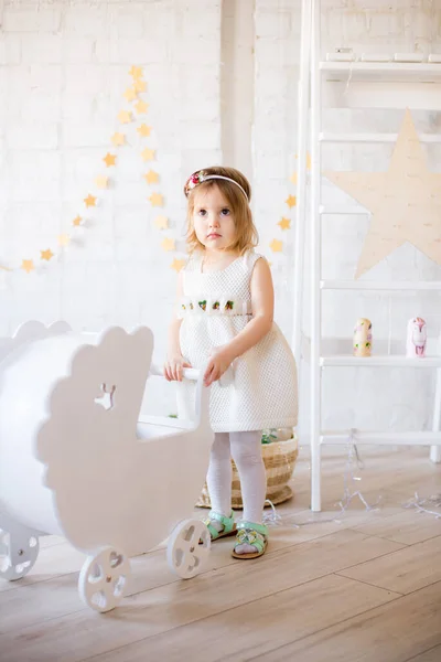白いドレスの小さなかわいい女の子は おもちゃやガーランドで飾られた明るい子供部屋の人形のための白いペッグで遊ぶ — ストック写真