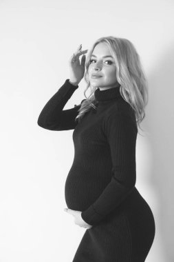 Stüdyoda koyu renk elbiseli, sarışın, genç ve güzel hamile bir kadın var. Hamile kadınlar için moda. Mutlu annelik