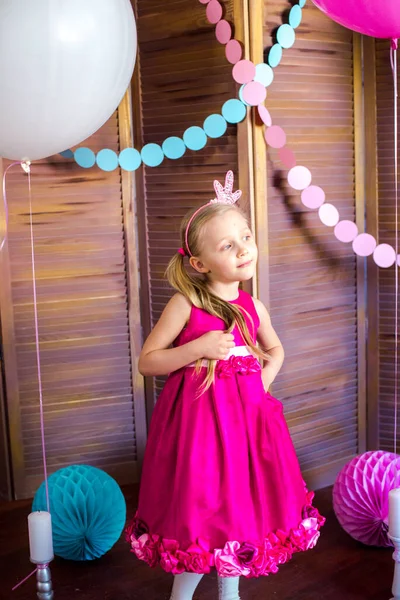 Little cute girl with blond hair in a pink dress and a princess crown with large bright balloons and garlands. Children's holiday. Children's decor. Balloons