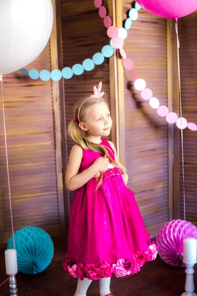 Little cute girl with blond hair in a pink dress and a princess crown with large bright balloons and garlands. Children's holiday. Children's decor. Balloons