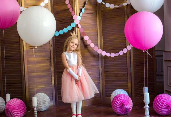 Little cute girl with blond hair in a pink dress and a princess crown with large bright balloons and garlands. Children's holiday. Children's decor. Balloons