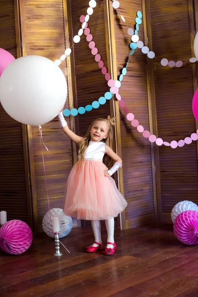 Little cute girl with blond hair in a pink dress and a princess crown with large bright balloons and garlands. Children\'s holiday. Children\'s decor. Balloons