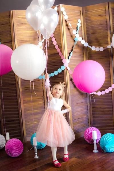 Little cute girl with blond hair in a pink dress and a princess crown with large bright balloons and garlands. Children's holiday. Children's decor. Balloons