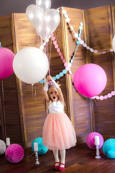 Little cute girl with blond hair in a pink dress and a princess crown with large bright balloons and garlands. Children\'s holiday. Children\'s decor. Balloons