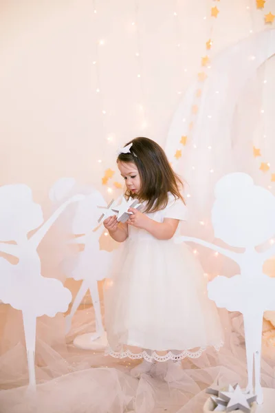 穿着白色公主礼服 头发上戴着蝴蝶结的可爱小女孩 在金星般的星空中 在一个巨大的白色木制月亮和花环的衬托下 与木制芭蕾舞演员嬉戏 儿童的装饰 儿童假日 — 图库照片