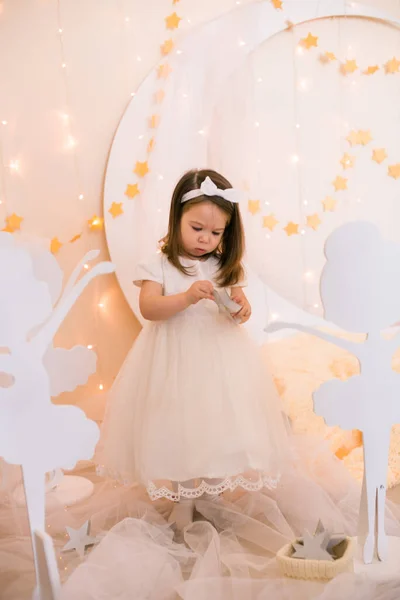 穿着白色公主礼服 头发上戴着蝴蝶结的可爱小女孩 在金星般的星空中 在一个巨大的白色木制月亮和花环的衬托下 与木制芭蕾舞演员嬉戏 儿童的装饰 儿童假日 — 图库照片