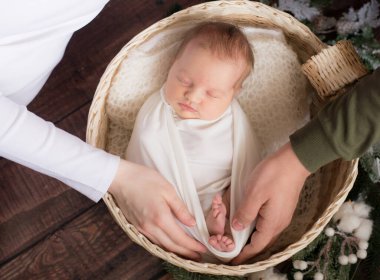 Anne ve babanın elleri, ahşap zeminde ağaç dalları ve pamukla süslenmiş hasır bir sepetin içinde beyaz bir battaniyede şirin bebek bacakları tutuyor. Mutlu annelik
