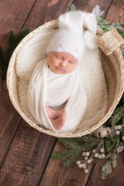 Beyaz battaniyeli şirin bir bebek ve ahşap zeminde iğne dalları ve pamukla süslenmiş hasır bir sepetin içinde beyaz örülmüş bir şapka. Mutlu annelik