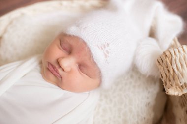 Beyaz battaniyeli şirin bir bebek ve ahşap zeminde iğne dalları ve pamukla süslenmiş hasır bir sepetin içinde beyaz örülmüş bir şapka. Mutlu annelik