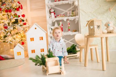 Sarı saçlı şirin çocuk, Noel ağacının yanında süslenmiş parlak bir odada ahşap eko oyuncaklarla oynuyor. Mutlu bir çocukluk. Noel havası