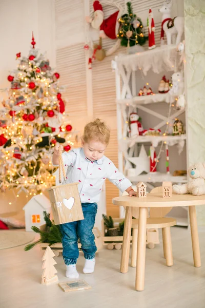 Sarı saçlı şirin çocuk, Noel ağacının yanında süslenmiş parlak bir odada ahşap eko oyuncaklarla oynuyor. Mutlu bir çocukluk. Noel havası