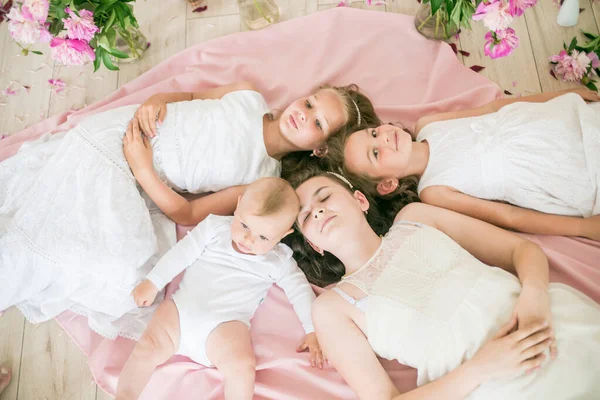 可爱的孩子们 一个身穿白衣的弟弟和姐姐们一起躺在一个明亮的春天工作室的地板上 房间里有粉色的牡丹 快乐的家庭快乐的童年 — 图库照片
