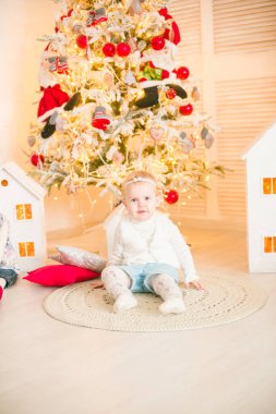 Sarı saçlı şirin küçük kız, Noel ağacının yanındaki parlak bir odada Noel çelenkleriyle oynuyor. Mutlu bir çocukluk. Noel havası