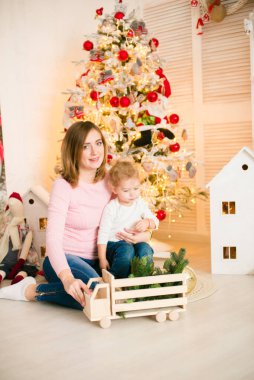 Sarışın, sevimli küçük çocuk, Noel ağacının yanında süslenmiş parlak bir odada annesiyle oynuyor. Mutlu bir çocukluk. Noel havası