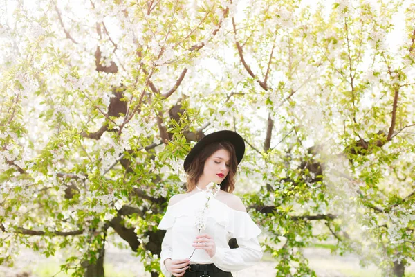 美丽的小女孩 戴着黑色的帽子 穿着白色的浪漫的丝绸衬衫 在春天里的一个盛开的樱桃园里 手里拿着盛开的樱花枝 春天与时尚 — 图库照片