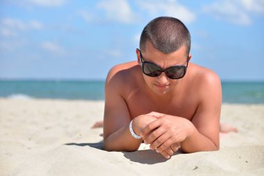 Sahilde güneş gözlüğü takan bir adam kumda yan bakıyor. Yazgan