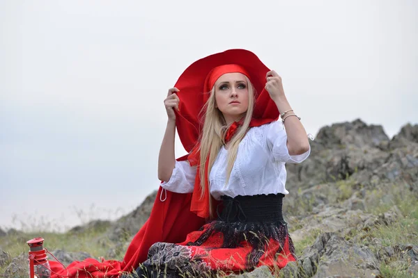 Beautiful blonde woman with red cloak sitting on a rock on the e — Stock Photo, Image