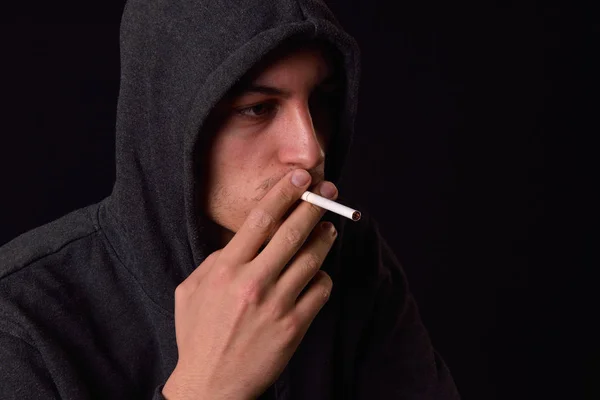 Teenager in a black hooded sweatshirt smoking a cigarette on a d — Stock Photo, Image