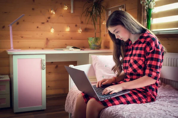 Young schoolgirl learns lessons at a distance at home in a cozy wooden interior on the bed.