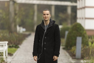 Genç adam gülümsüyor ve kameraya bakıyor. Kapıda durup kulaklık dinliyor..