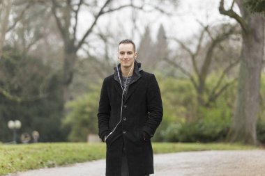 Parkta gülümseyen, palto giyen ve kulaklık dinleyen genç bir adam. Orta boy..