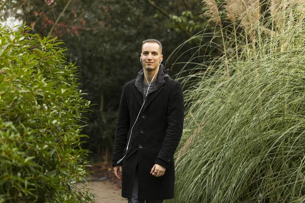 Homme caucasien avec écouteurs et manteau noir debout dans le jardin décoratif — Photo