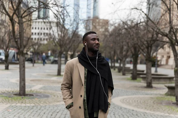 Fashionable young black man with earphones and beige coat standing in city and looking away.