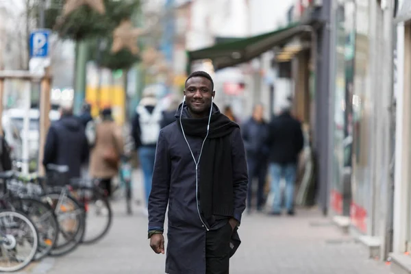 Young black man with earphones and long coat walking towards camera on street.
