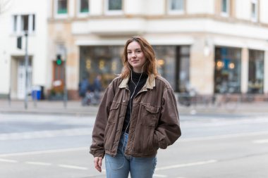 Kulaklık dinleyen, kahverengi ceketli, kameraya gülümseyen beyaz bir kadın..