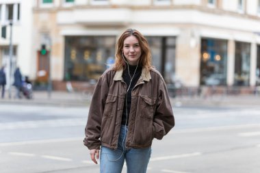 Şehirde kot pantolon ve ceket giyen ve kameraya gülümseyen kulaklıkları dinleyen genç bir kadın..