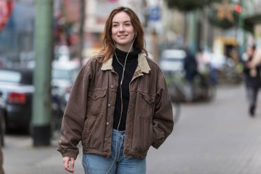 Sonbaharda kaldırımda dişleri pürüzsüz bir şekilde kulaklık dinleyen genç bir kadın. Uzaklara bakıyorum..