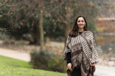 Genç orta doğulu kadın panço giyiyor ve parkta gülümsüyor..