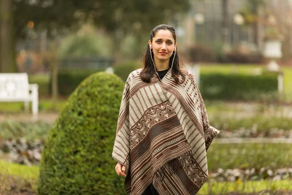 Female Indian model dressed in poncho posing outdoors in ornamental garden. Front view.