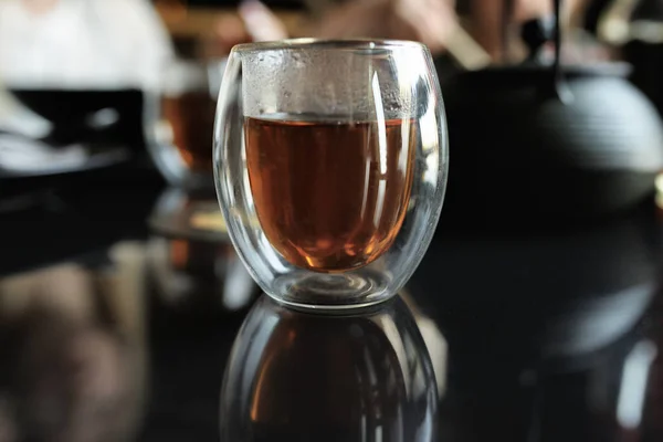 on a dark mirror table is a glass of tea, a teapot on the background in defocus