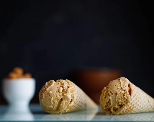 two caramel ice cream in waffle cones on a white glass surface on a black background, next to white coffee cups with caramel candies
