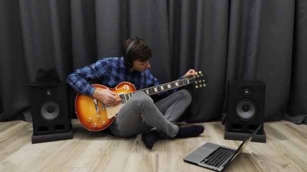 人間のチューニングエレキギター。リビングルームに座っている間、ヘッドフォンの男はノートブックを使用してギターをチューニングします。ギターのレッスンの準備をする若いプロのギタリスト — ストック動画