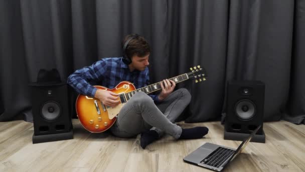 Human learning to play on guitar. Man in headphones practicing to play on electric guitar. Young teenager playing on guitar while sitting on floor at living room — 图库视频影像