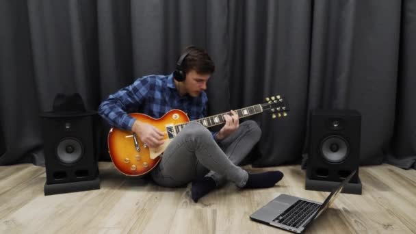 Hombre tocando en la guitarra eléctrica. Joven con auriculares profesionales y ropa casual practicando la guitarra usando un cuaderno. Adolescente cantando la canción en auriculares y tocando solo de guitarra — Vídeos de Stock